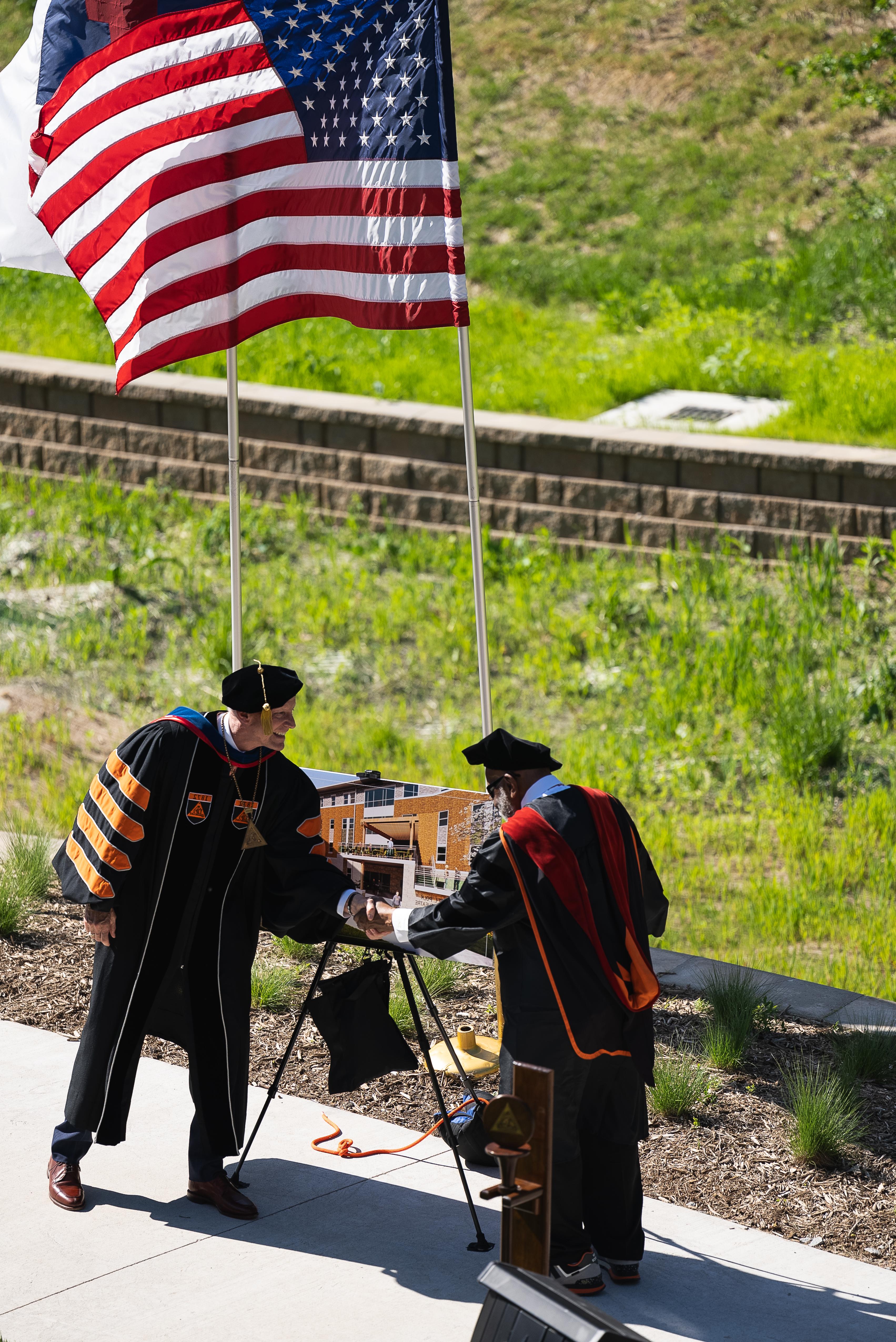 Two men in black academic robes shake hands in front of a poster showing a building. Behind them flies an American flag, with green plant growth in the background split by a brick retaining wall.